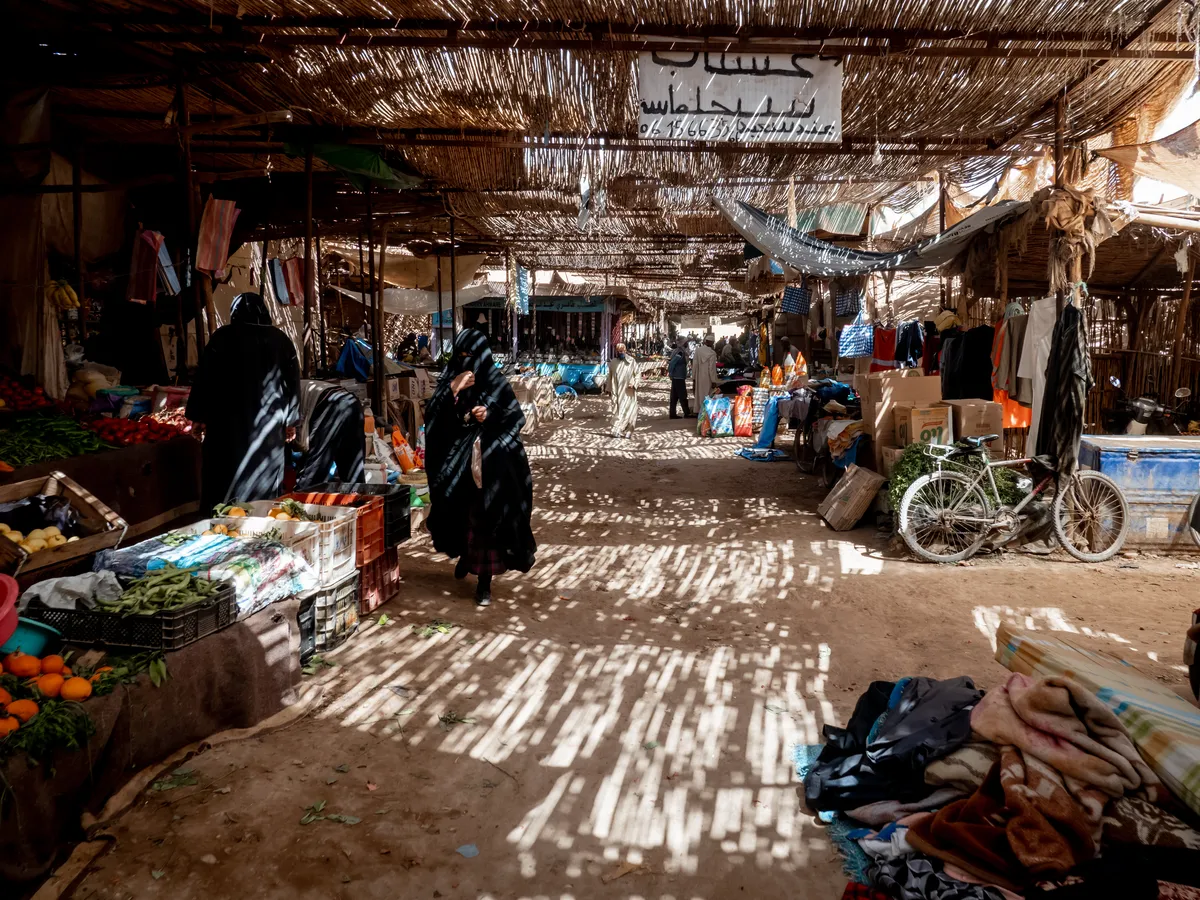 A covered market street, sunlight falling through the reed roof in stripes onto shoppers and stalls