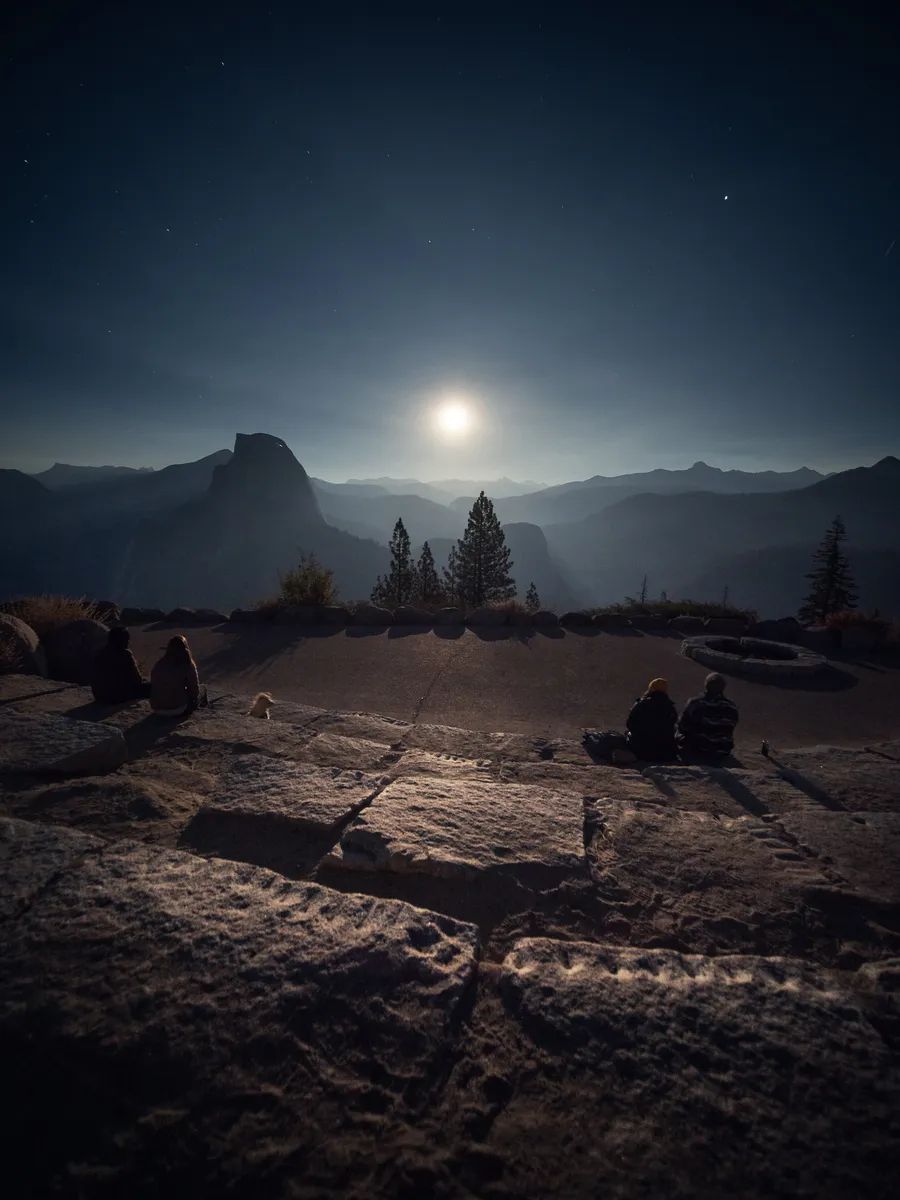 A full moon over Half Dome and the Yosemite valley, a few people sitting on the granite in the foreground